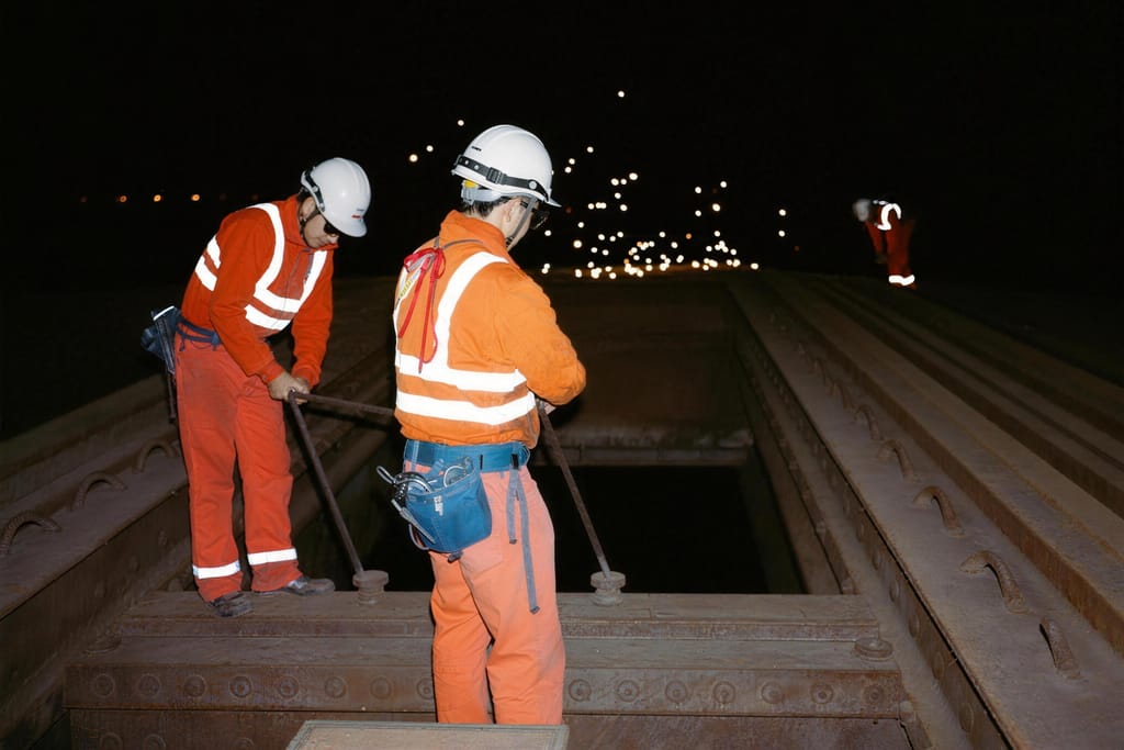 Two workers in high vis using long bars on a steel structure at night
