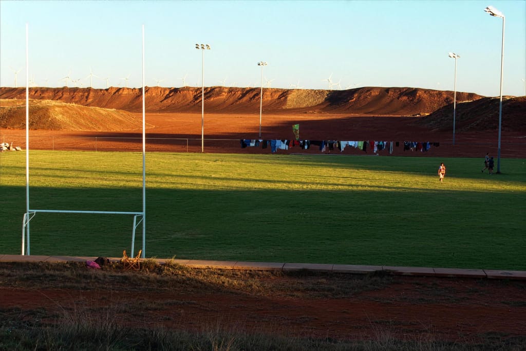 A grassed football oval with goal posts and light towers, red hills behind